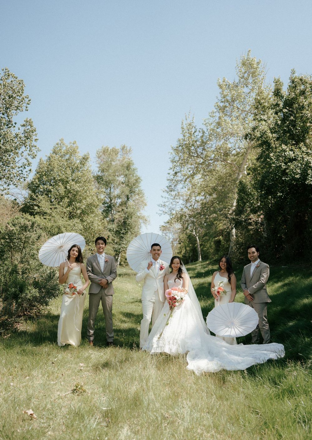 Joyful wedding party poses in a natural outdoor setting with green trees and sunlit grass.