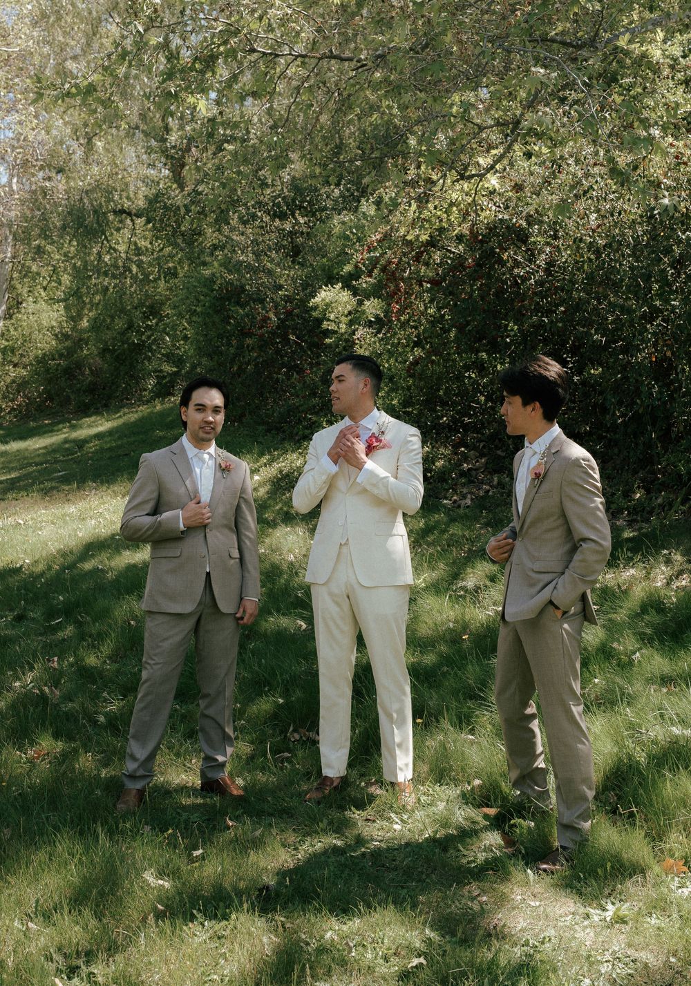 Three people in formal suits standing together on a grassy slope with trees in the background on a sunny day.