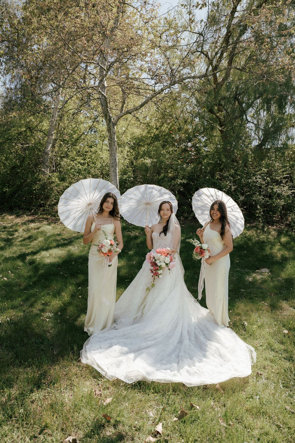 Three attendants in cream dresses pose with parasols beside a bride in white gown on a sunny hillside.