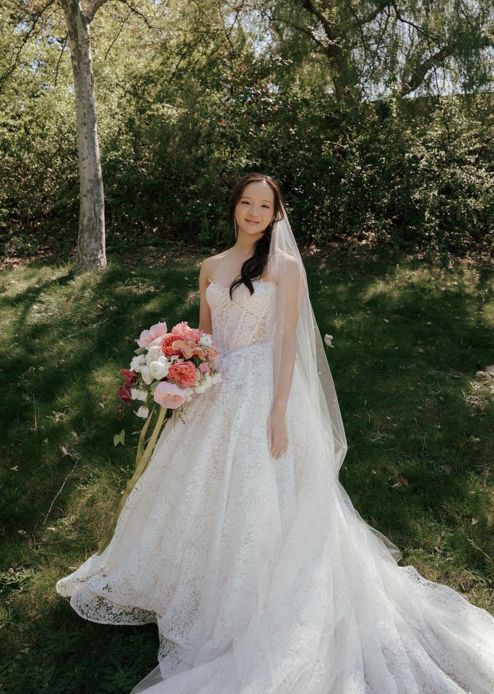 Elegant bride in a flowing white lace wedding gown stands in a sunlit garden holding a pink and coral bouquet.