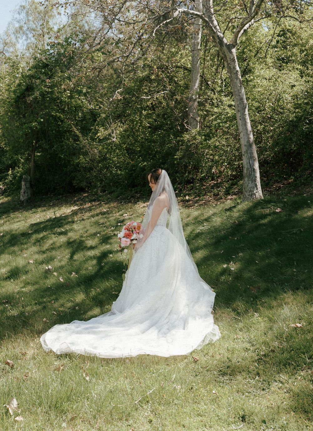 A wedding dress with flowing train cascades down a grassy hillside in dappled sunlight.