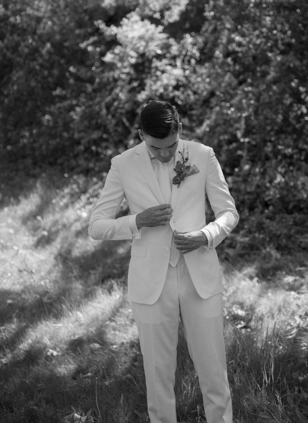 Black and white capture of wedding party members in light colored suits standing in sunlit grass.