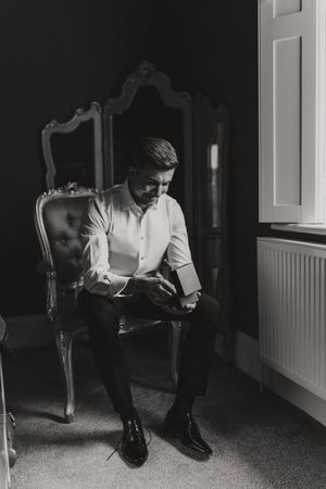 Black and white photo of a groom sitting in an ornate antique chair reading by window light at Hirst Priory - taken by Grimsby photographer.