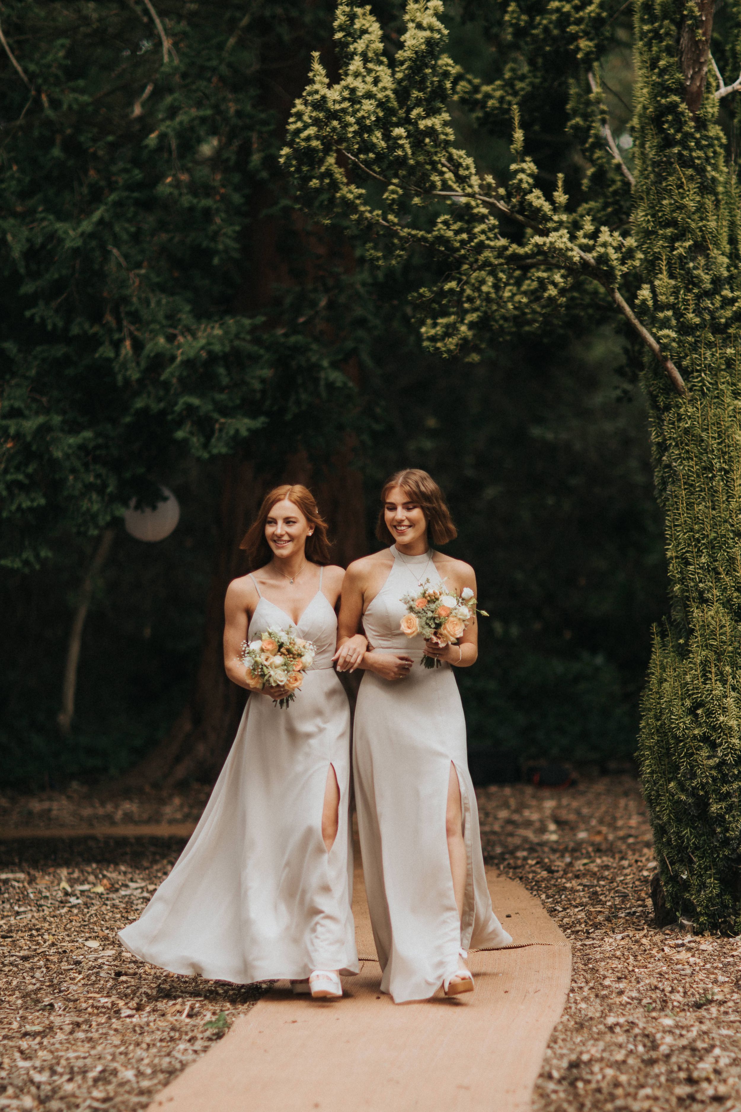 Two women in flowing white wedding dresses smile while standing on a forest path with autumn leaves at Hirst Priory.