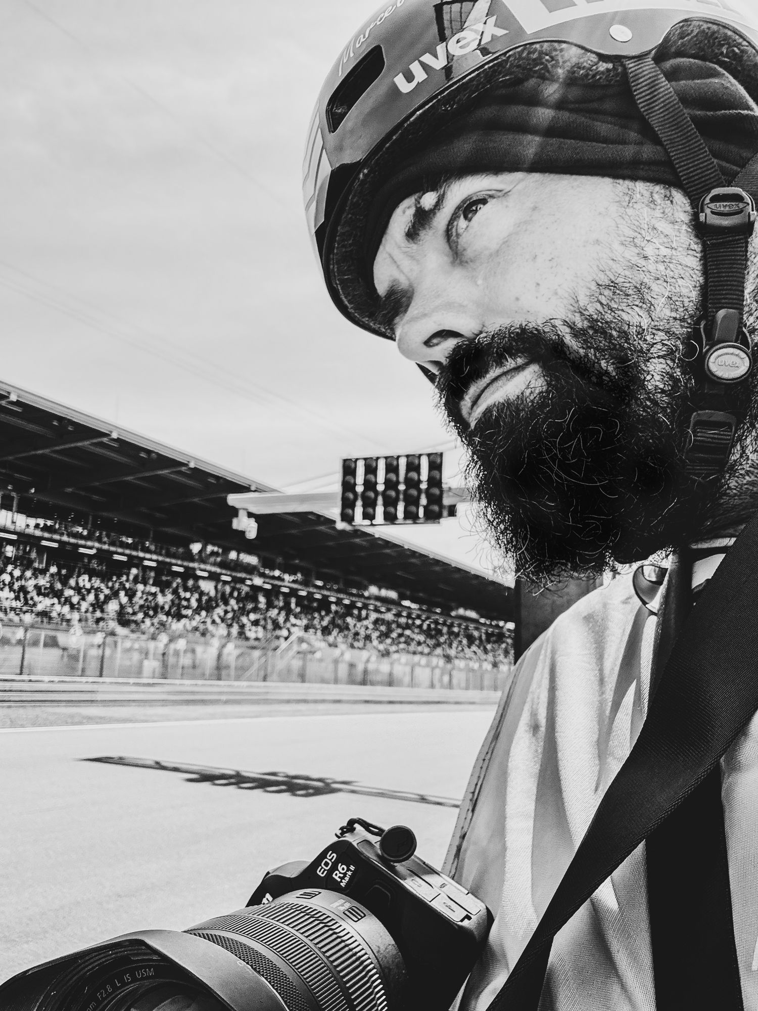 Black and white photo of a photographer with beard wearing helmet at a racetrack stadium.