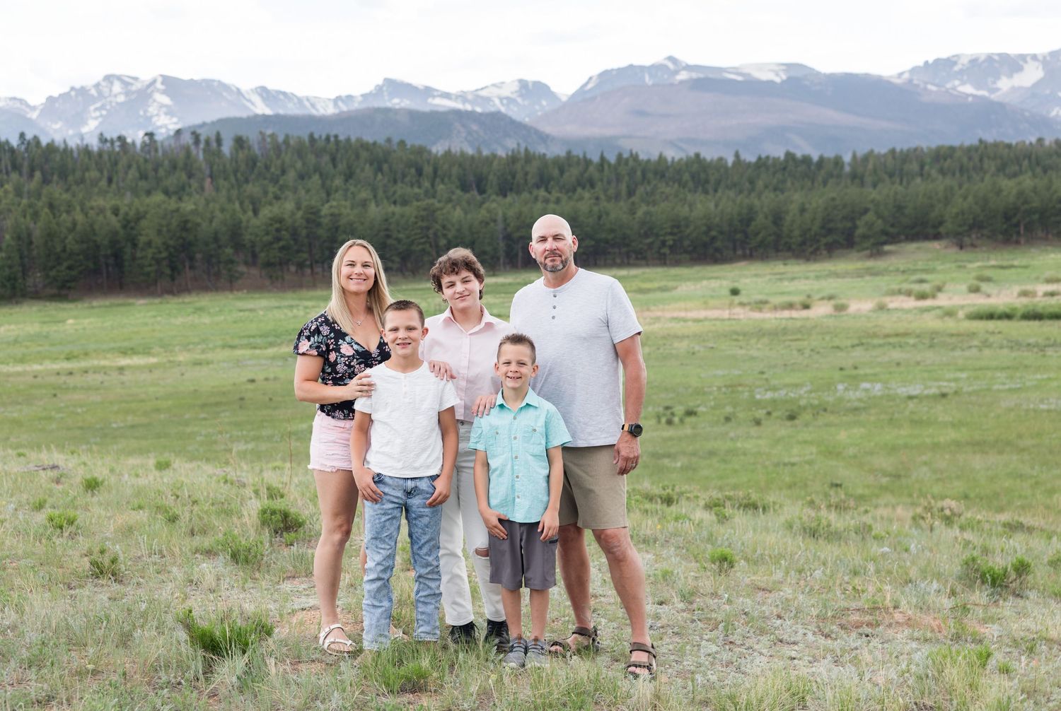 A group stands together in a meadow with snow-capped mountains and evergreen forest in the background.