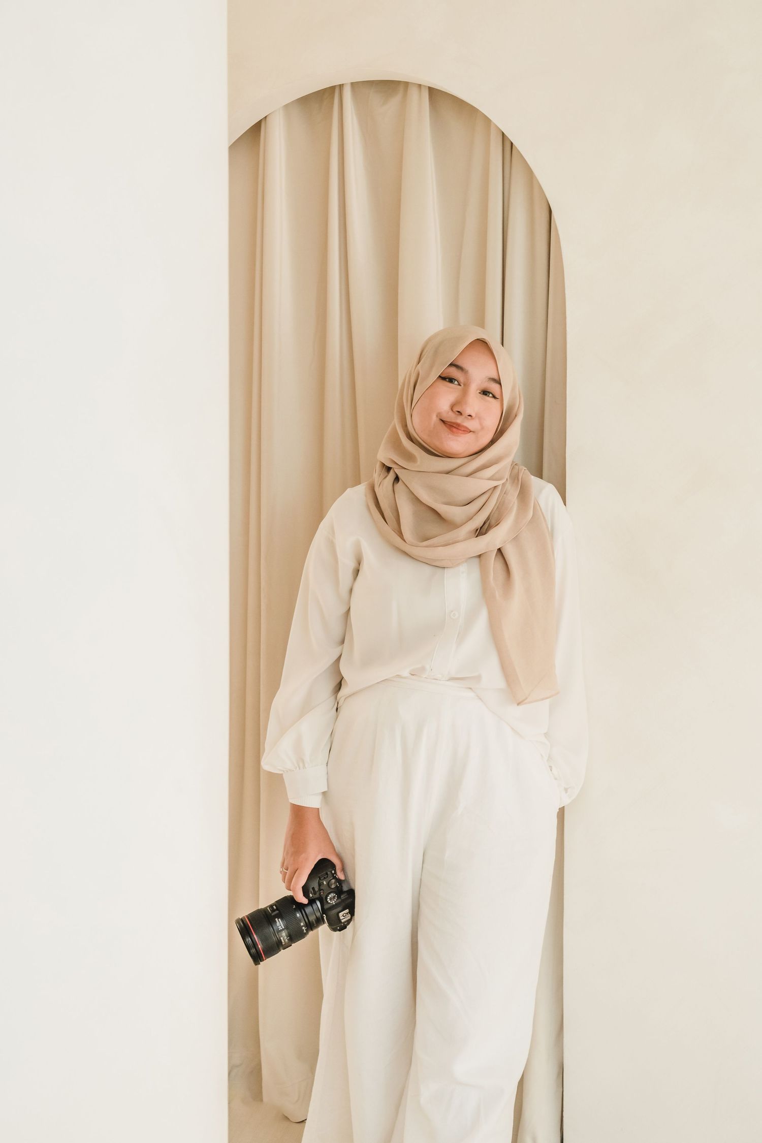 Person in cream hijab and white dress holding camera against neutral backdrop.