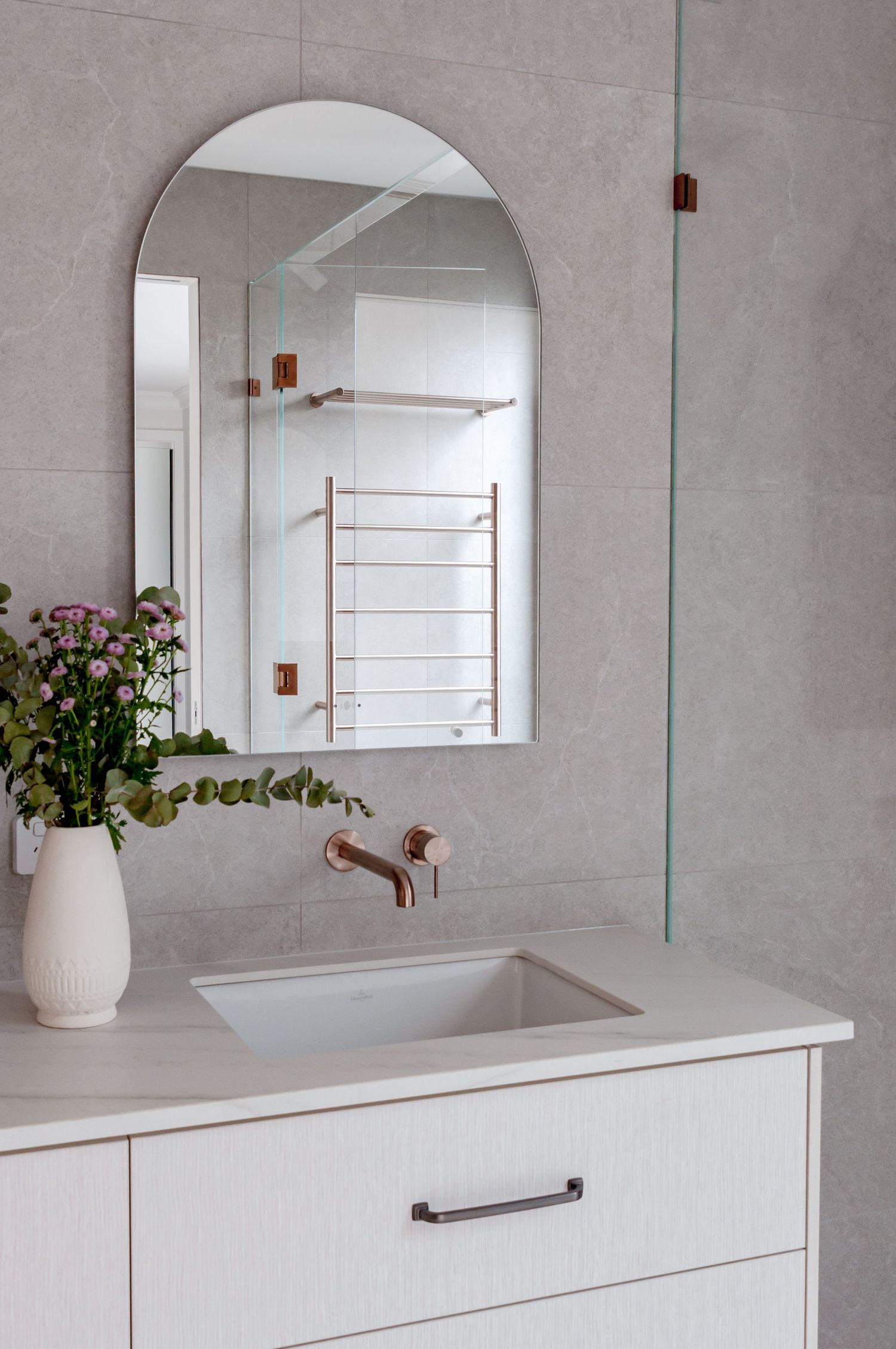 Elegant bathroom vanity with copper faucet, arched mirror, and white ceramic vase filled with pink flowers.