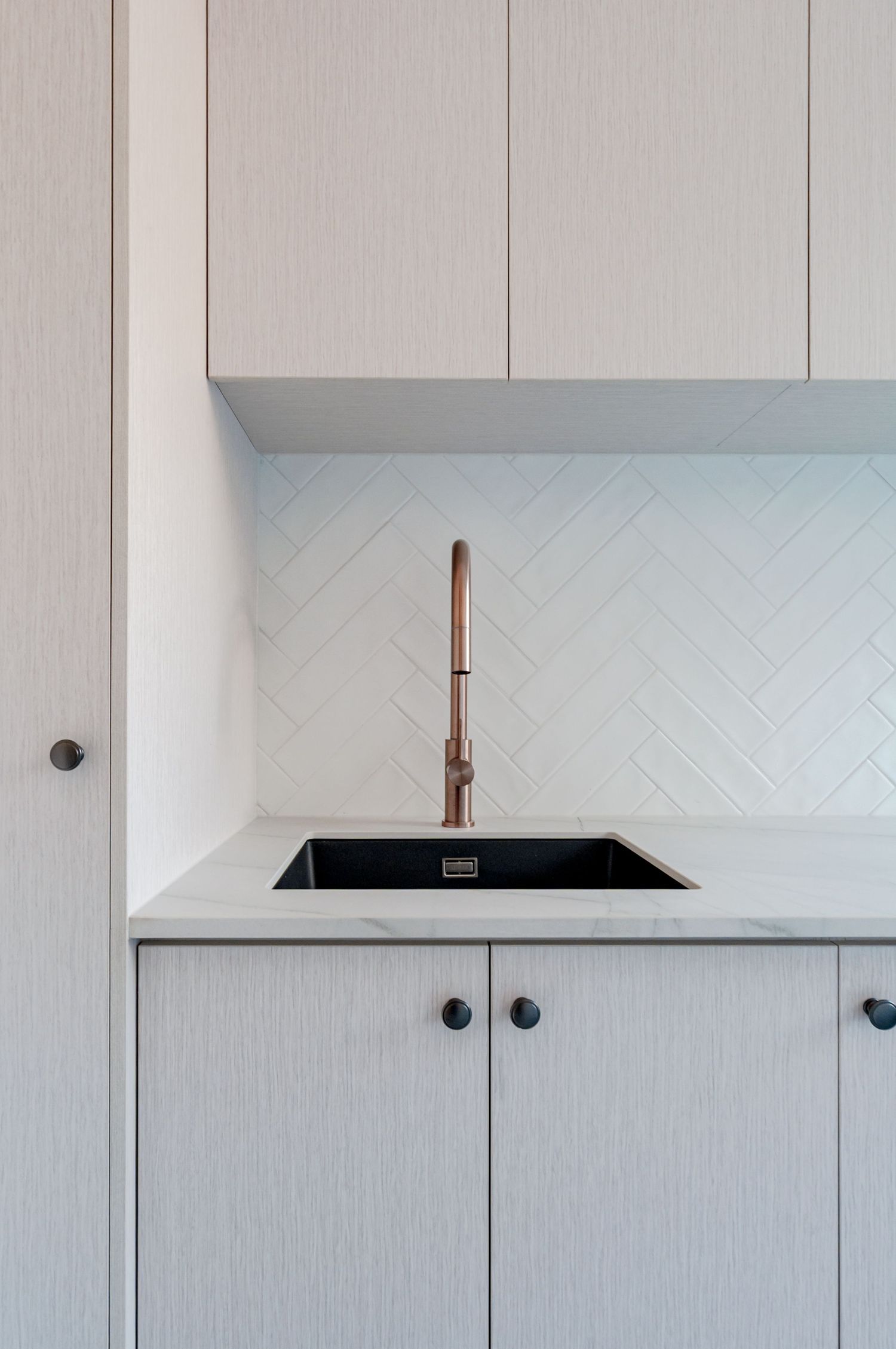Modern laundry sink with copper faucet against white herringbone tile backsplash and light wood cabinetry.