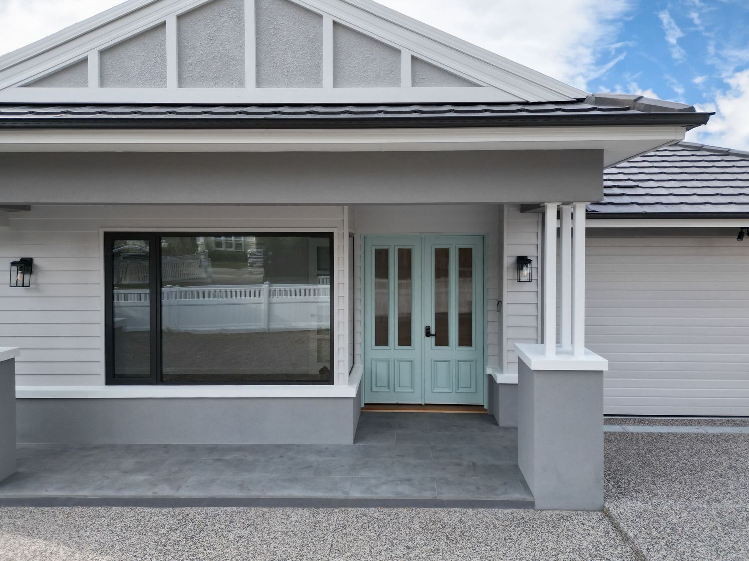 Modern home exterior featuring a light gray facade with teal front door and large picture window on concrete porch.
