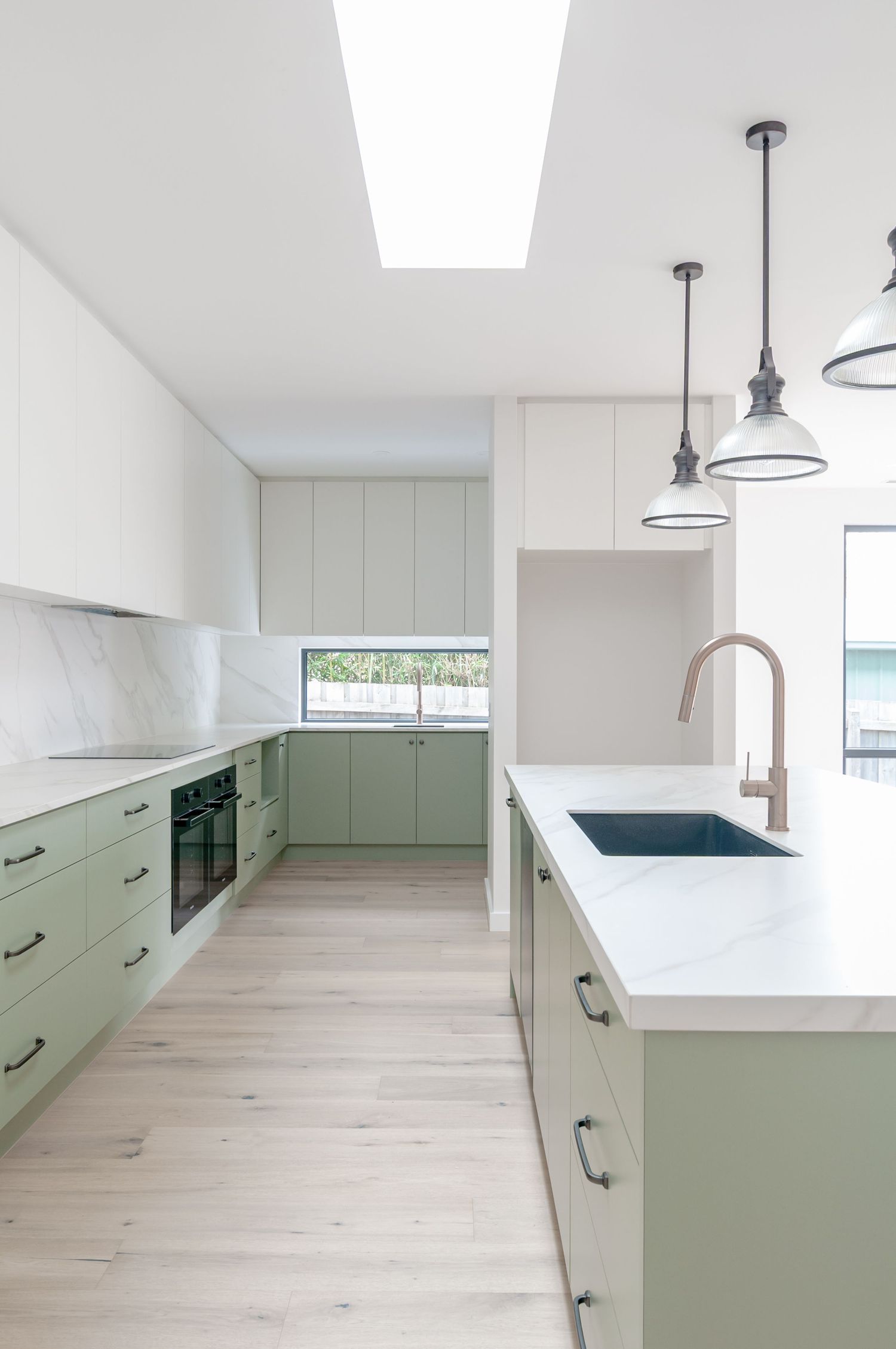Modern kitchen with sage green cabinets, white countertops, light wood floors and pendant lighting over white island.