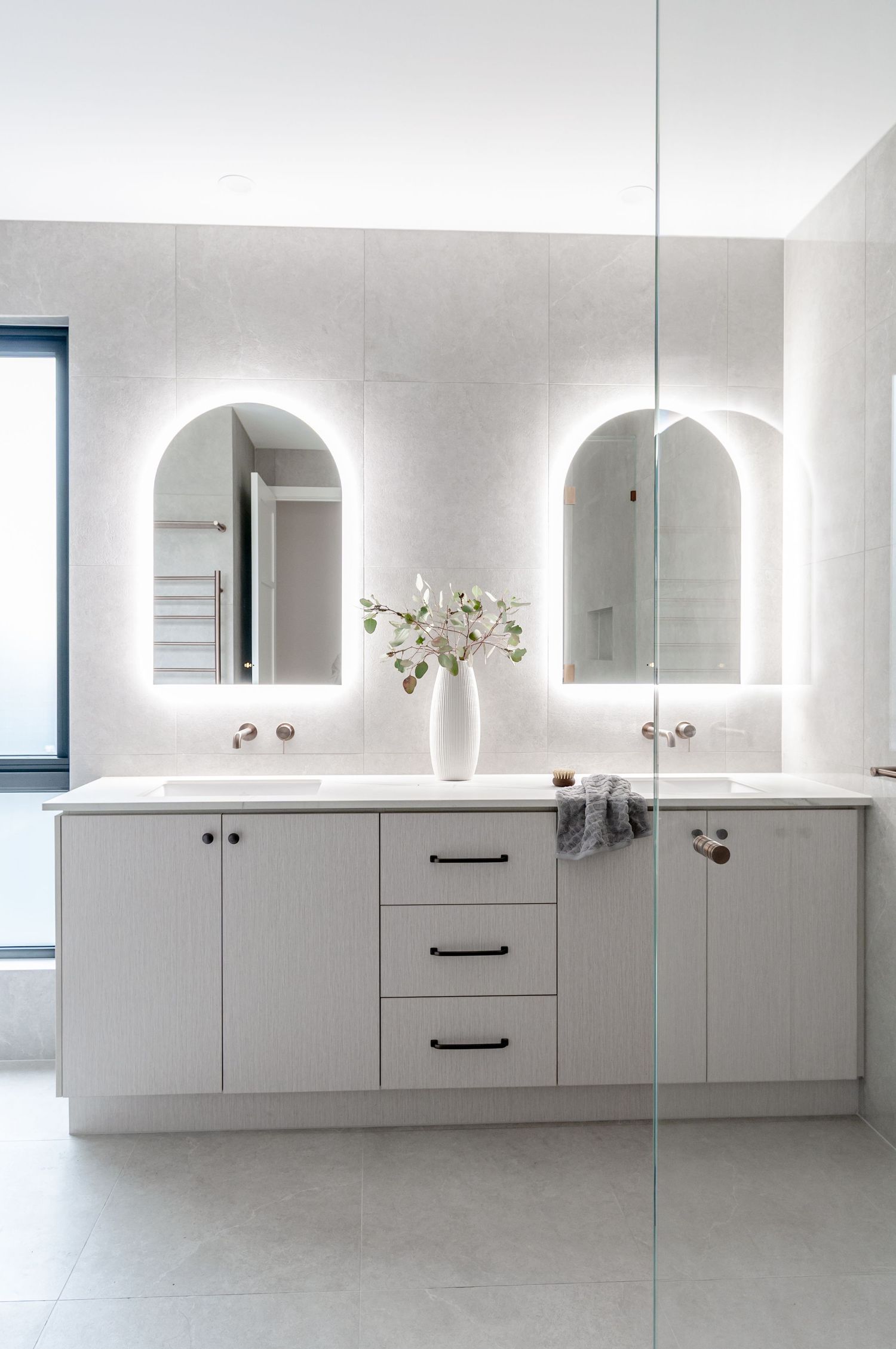 Modern white bathroom vanity with backlit arched mirrors, white vessel vase, and sleek cabinet hardware.