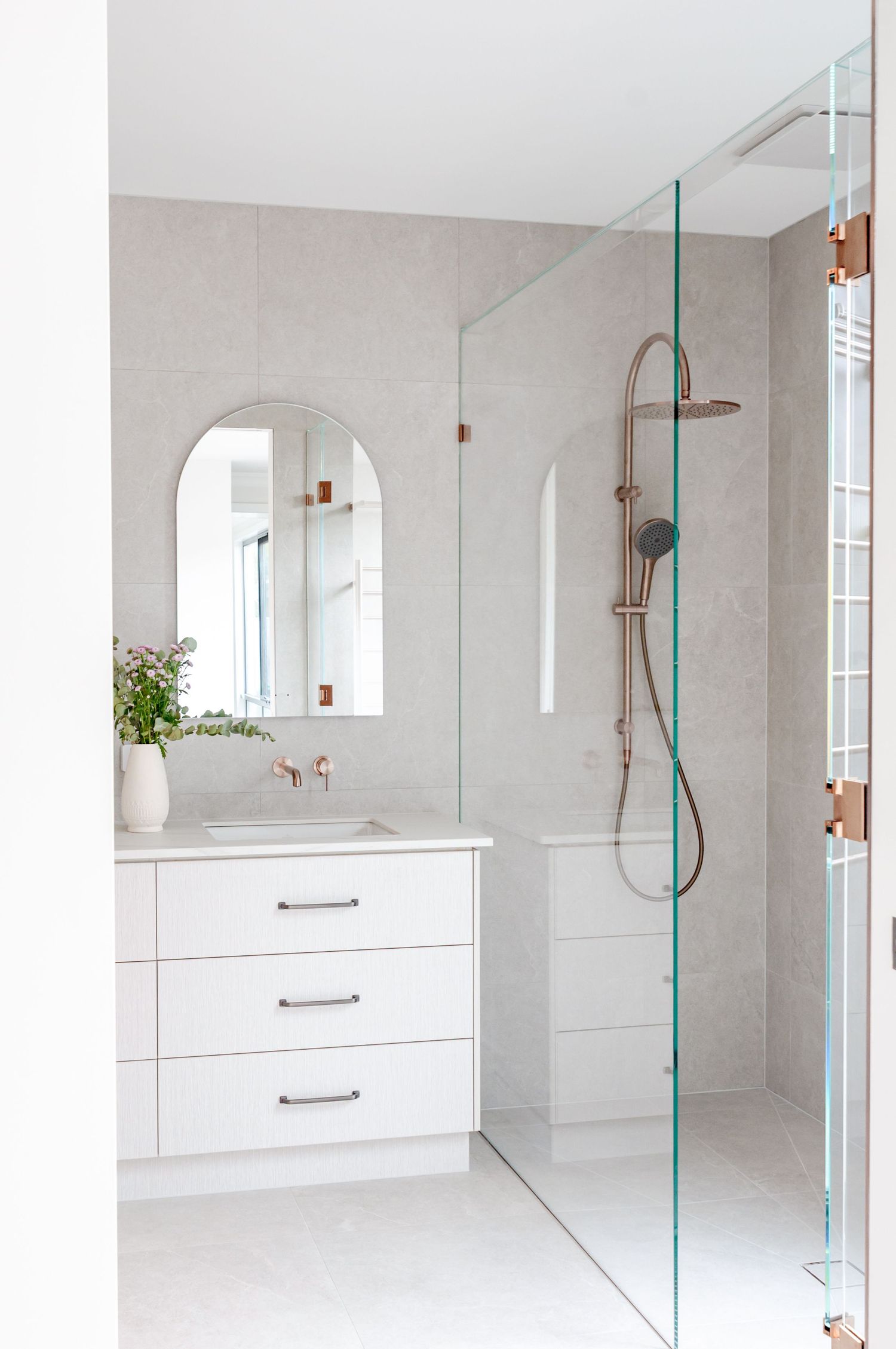 Modern white bathroom with glass shower partition, built-in vanity, and arched mirror niche in neutral tones.