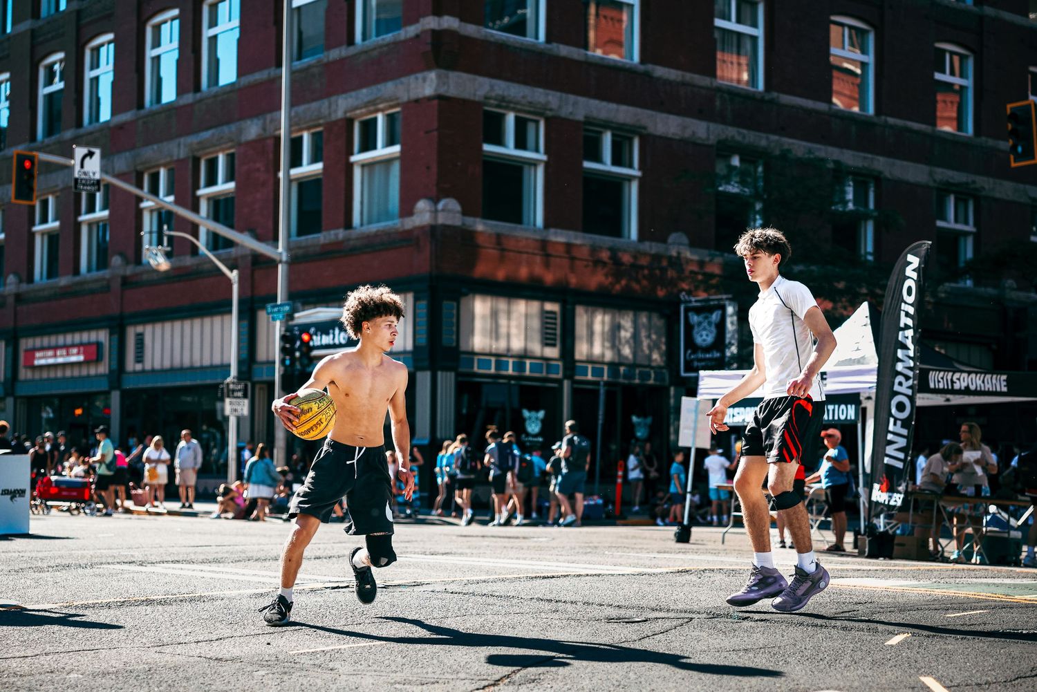 A person performs a skateboarding trick on a city street while a crowd watches in the background.