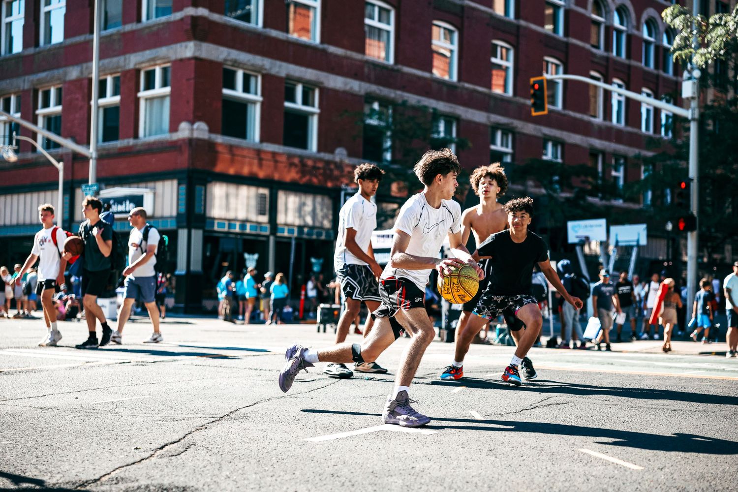 Athletes compete in an intense basketball game on an outdoor court surrounded by brick buildings and spectators.