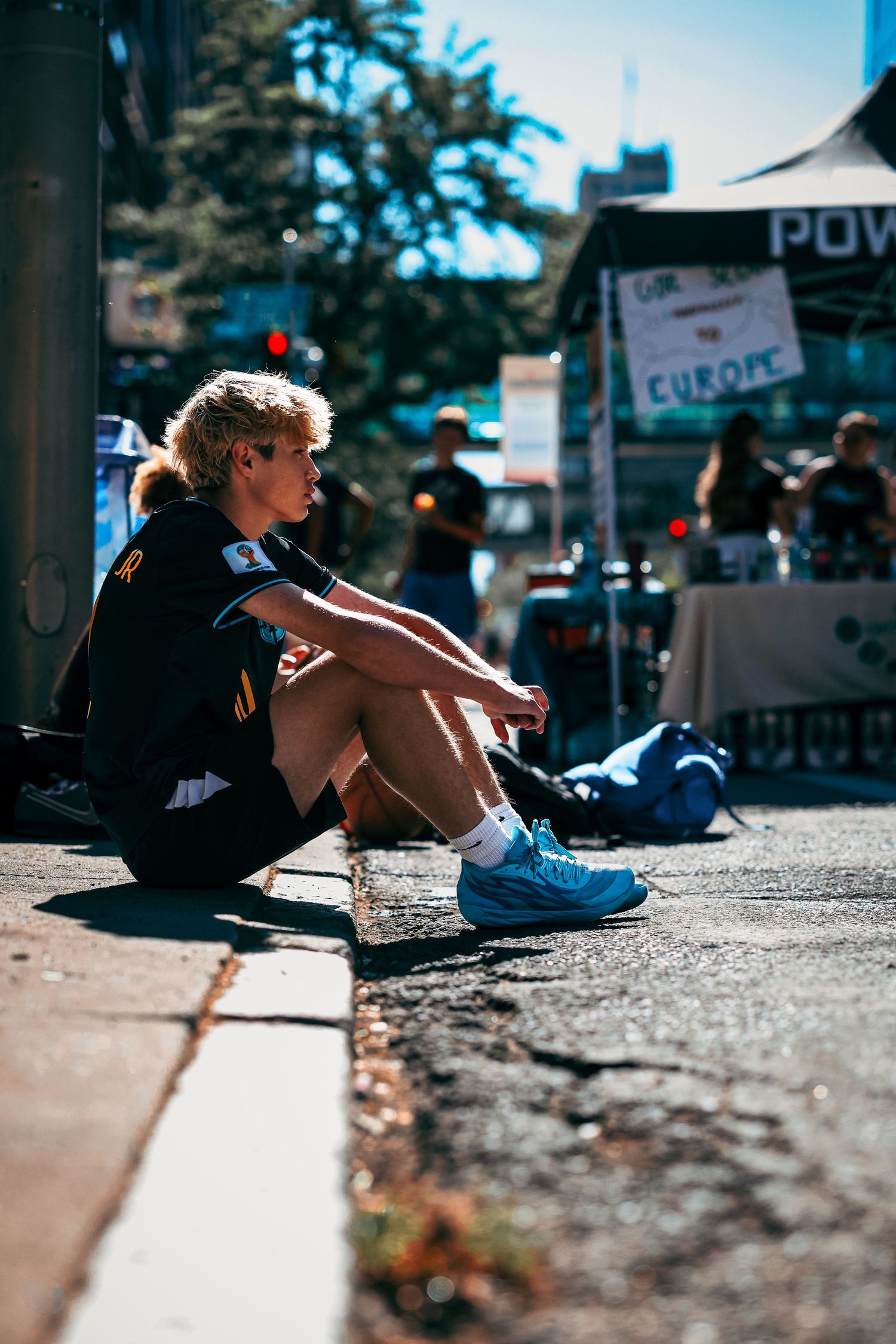 A person sits on a curb in an urban setting with blurred background elements on a sunny day.