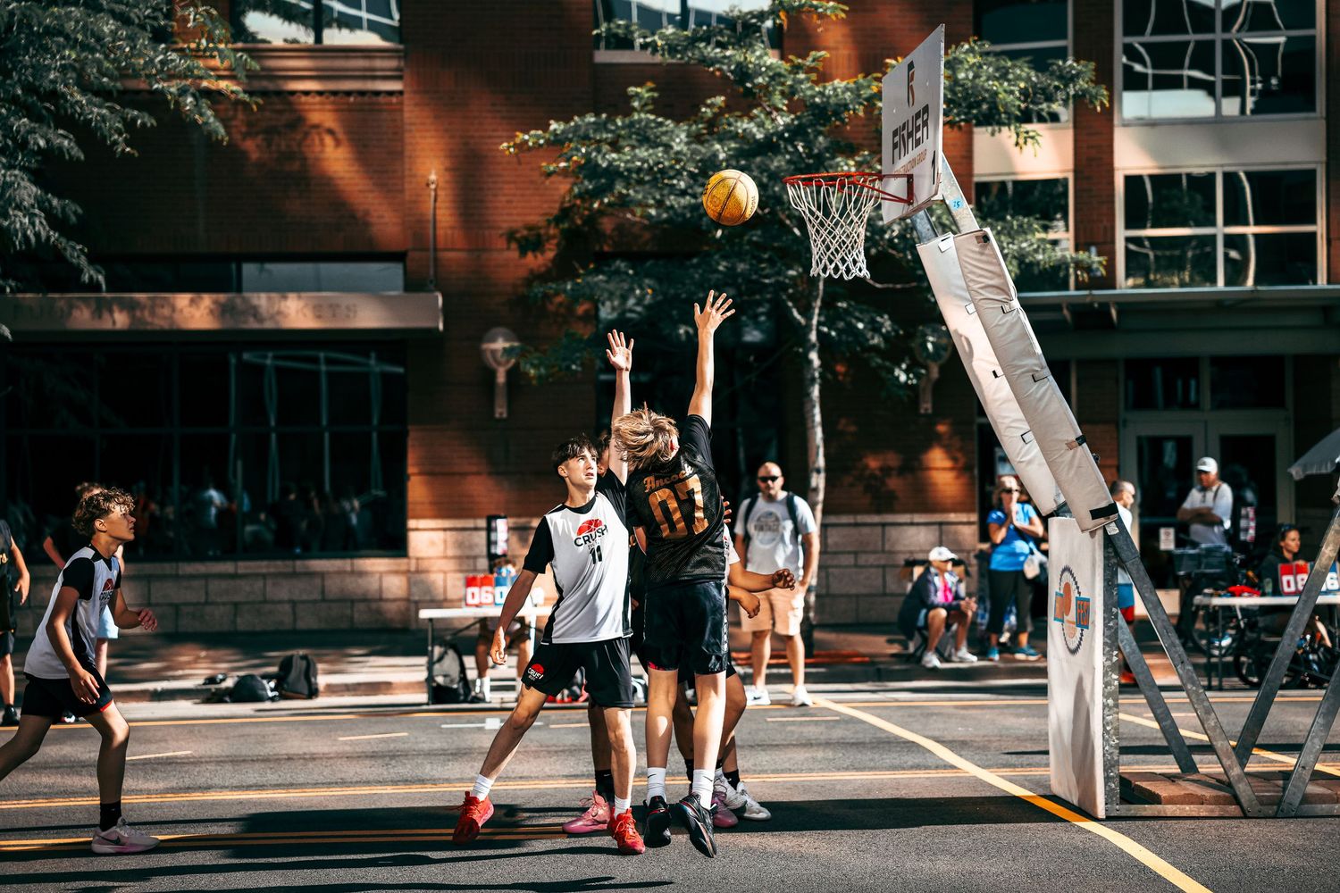 People playing basketball on an outdoor court with trees and buildings in the background.