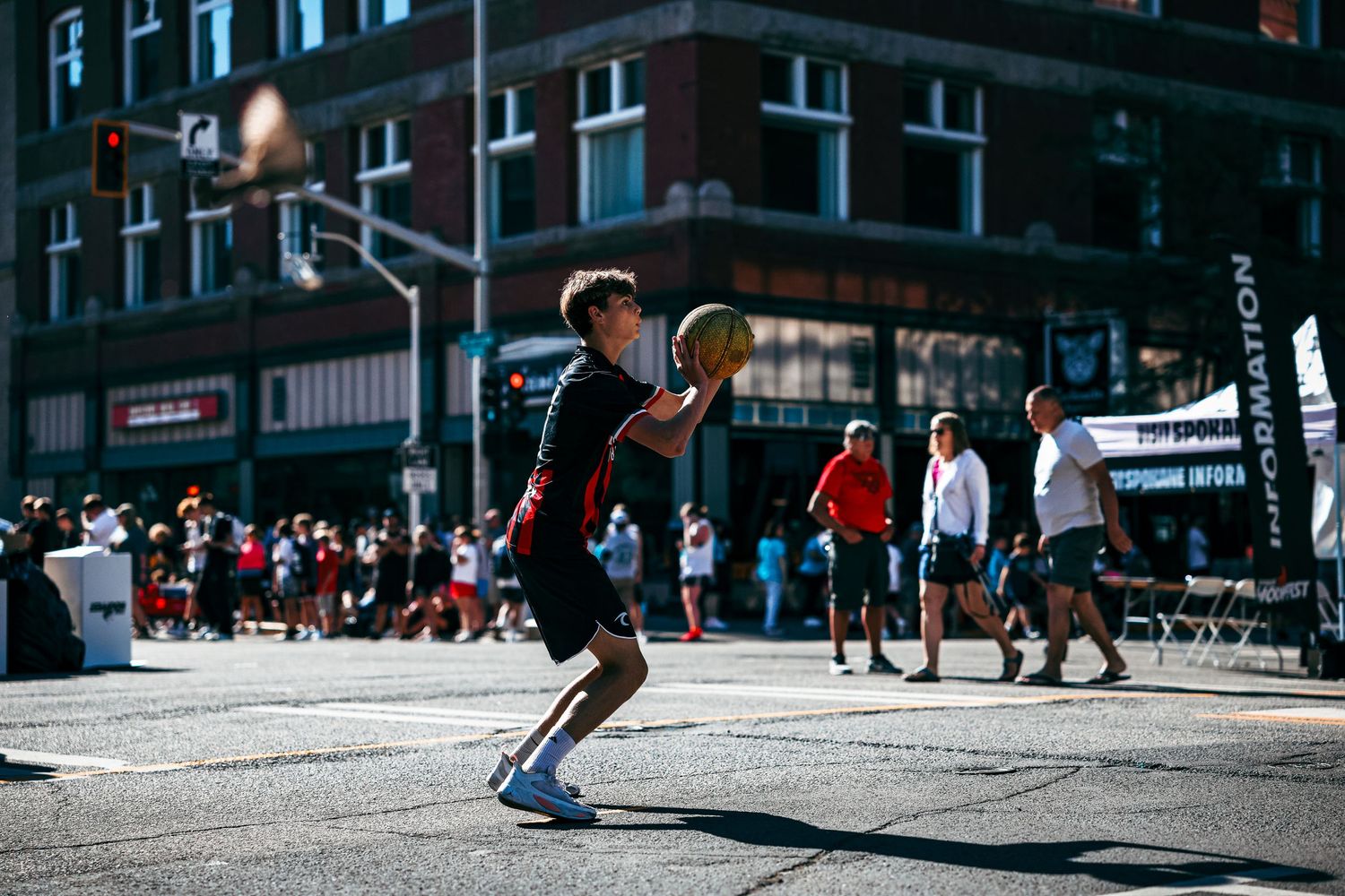 Basketball player in black jersey jumps to make a shot during a street game in an urban setting with spectators watching.