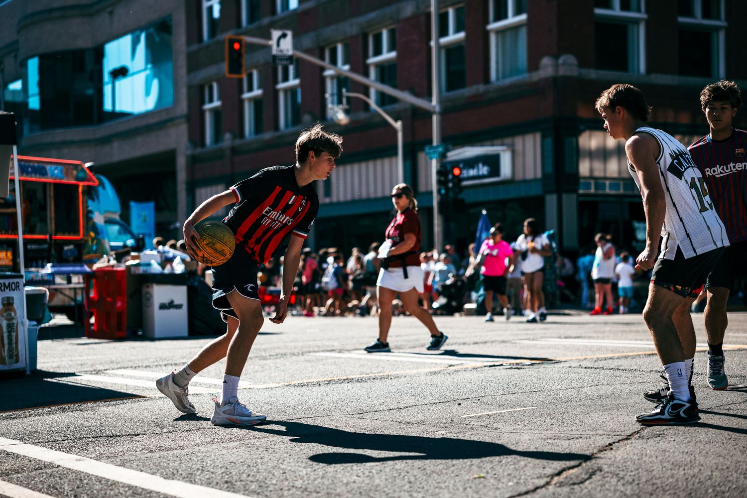 Athletes compete in an outdoor street basketball game on a sunny day in an urban setting with brick buildings in the background.