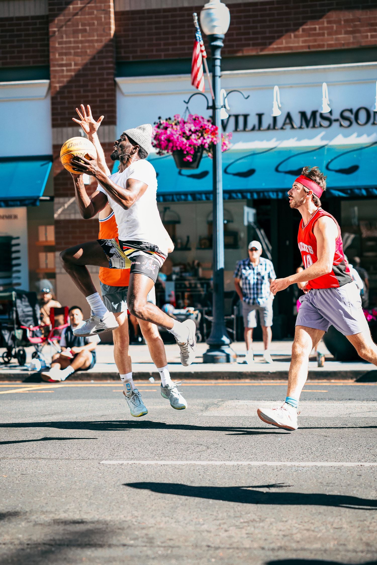 Two players leap during an intense street basketball game in front of a downtown storefront with hanging flower baskets.