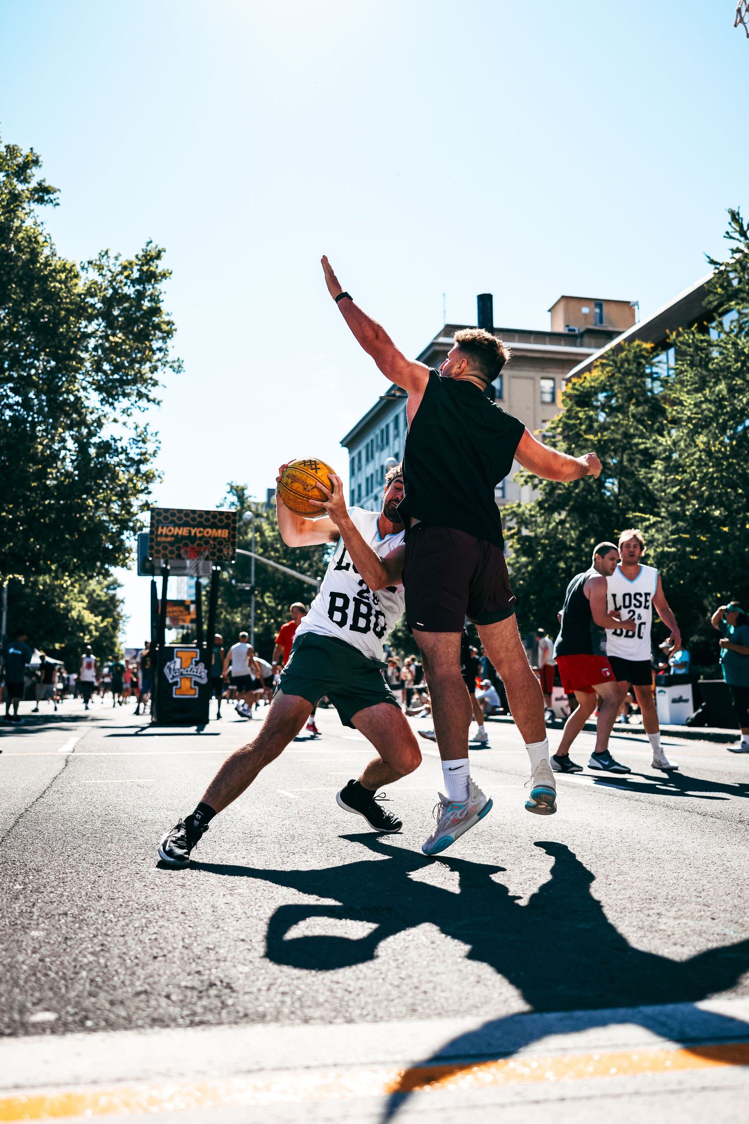 Urban street basketball players engage in dynamic gameplay with dramatic shadows cast on concrete court surface.