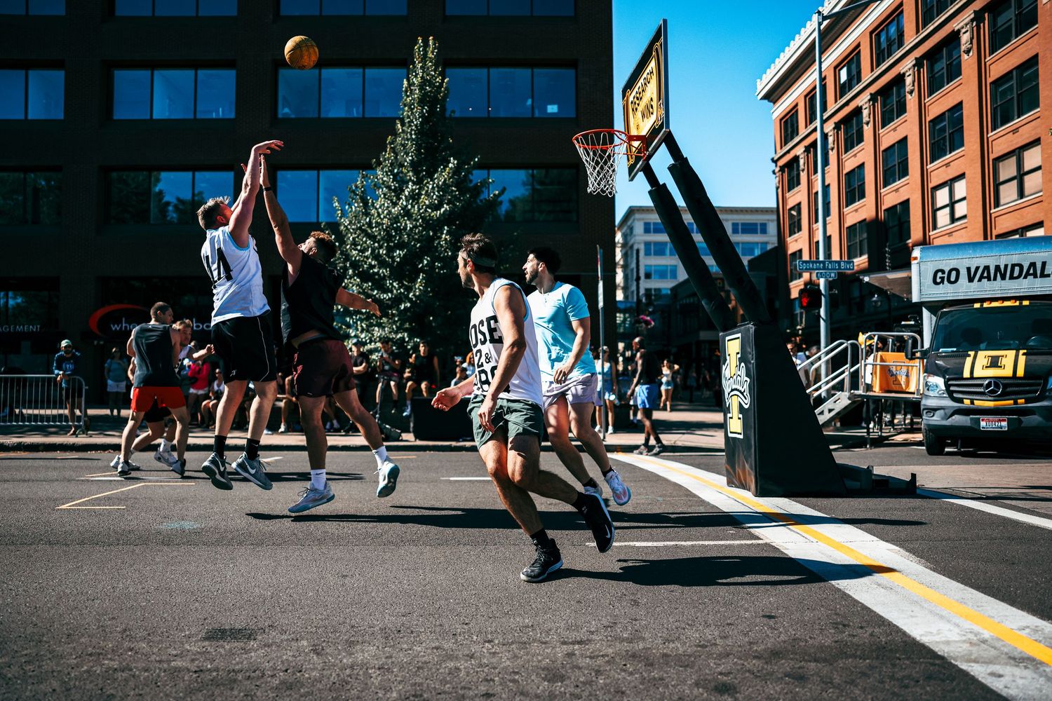 People playing street basketball on an urban court with tall buildings in the background during a sunny day.