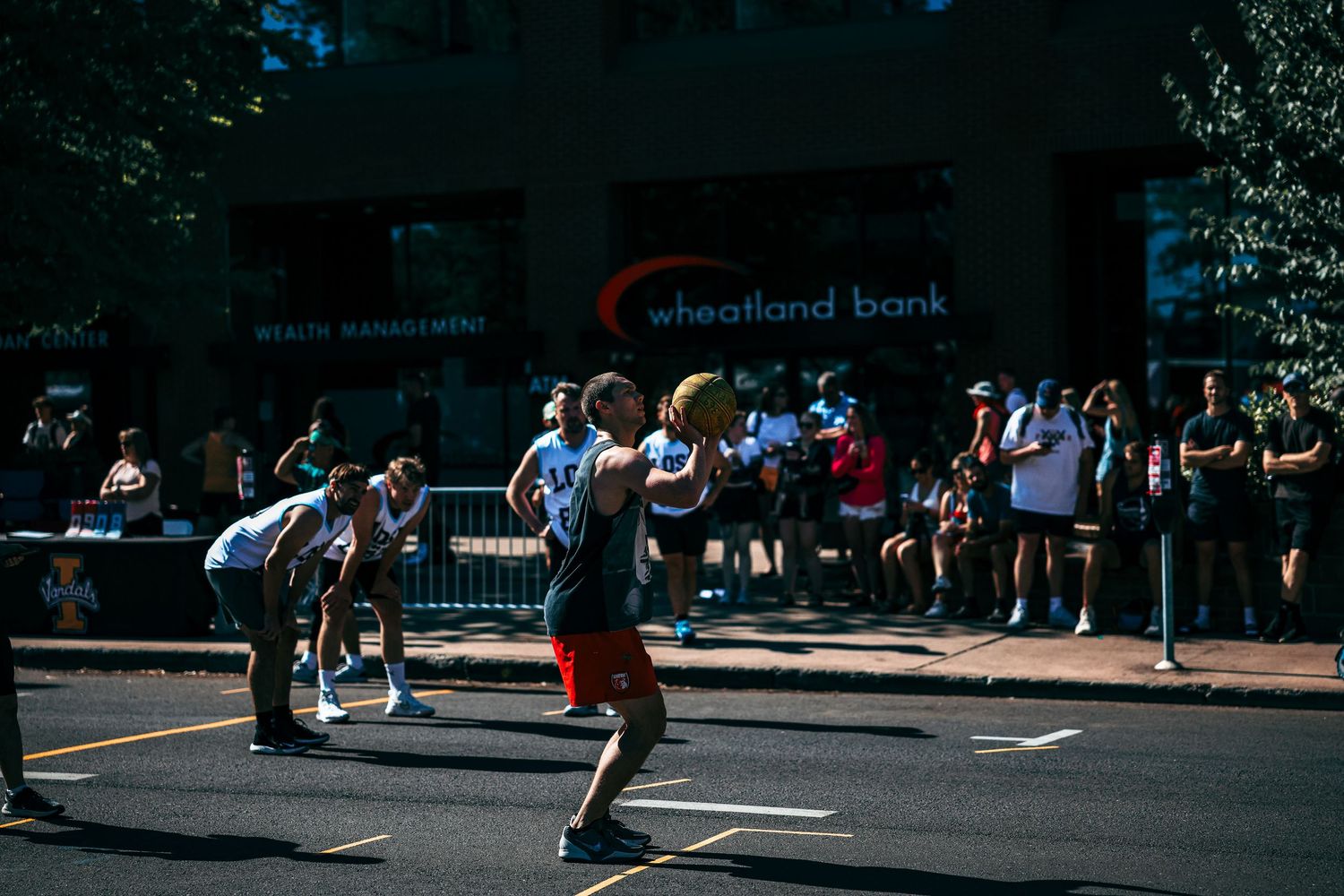 Runners pass by a crowd of spectators during a basketball tournament on a sunny day in the city.