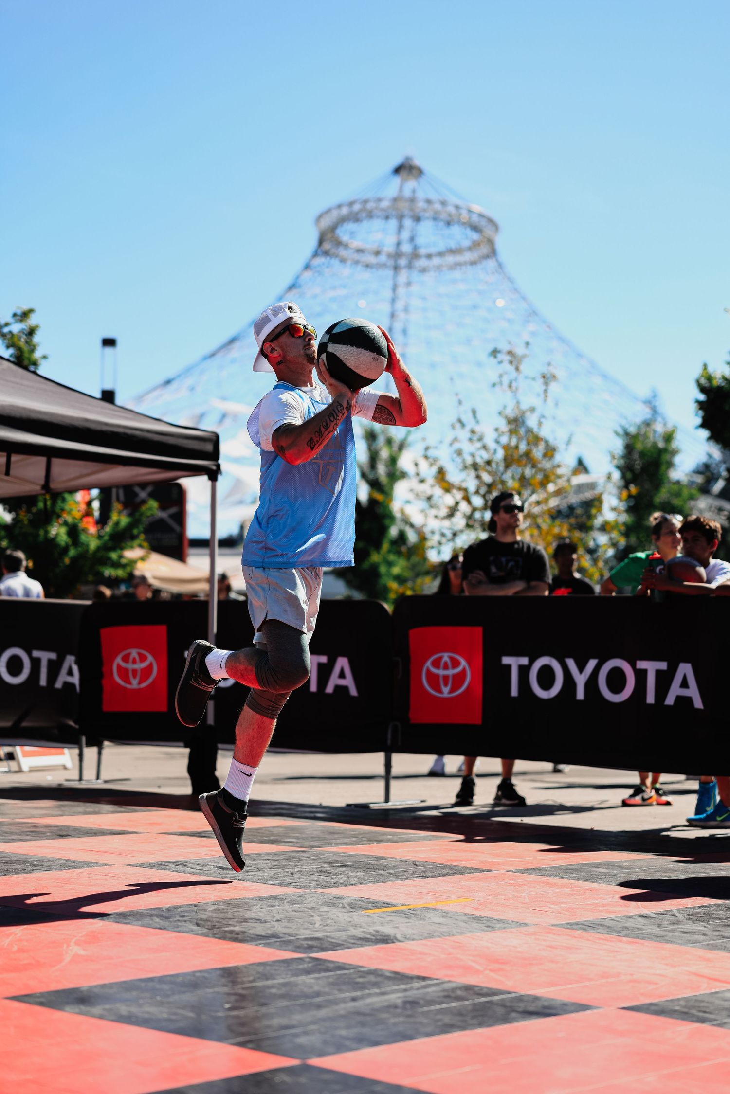 Basketball player jumps high during outdoor street game with Toyota banners and metal structure in background.