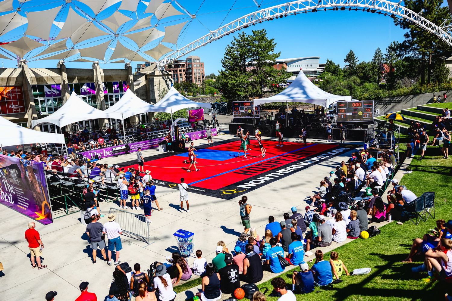 Large crowd gathers at outdoor event venue with white tents and red decorations during sunny summer day.