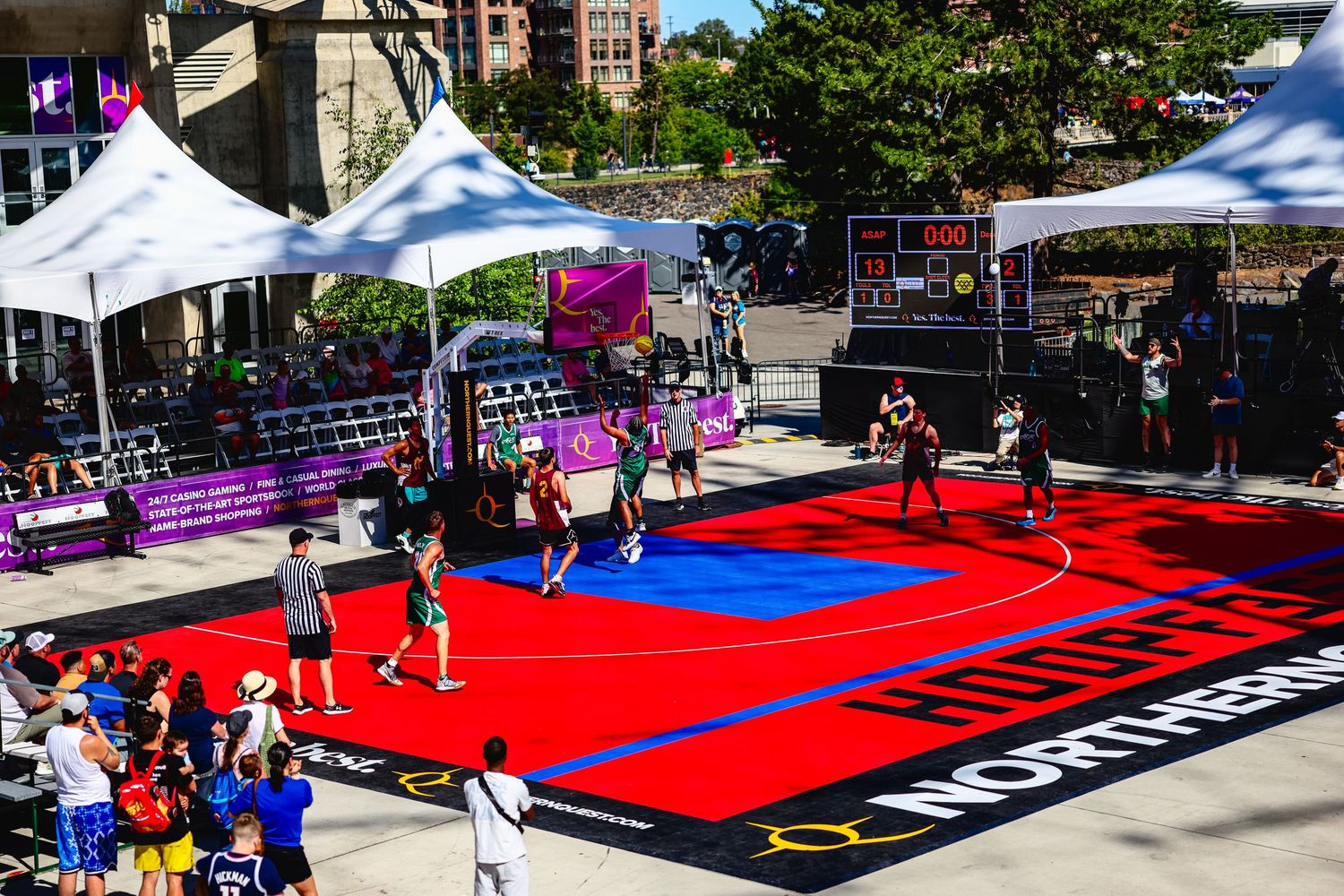 People play basketball on a vibrant red and blue outdoor court during a community event with white tents in the background.