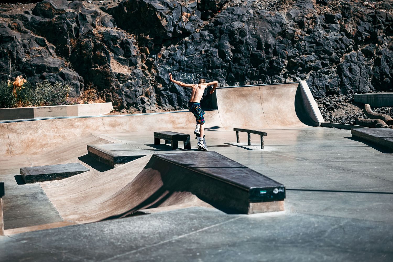 Concrete skate park ramps and obstacles against rocky cliff wall in outdoor setting.