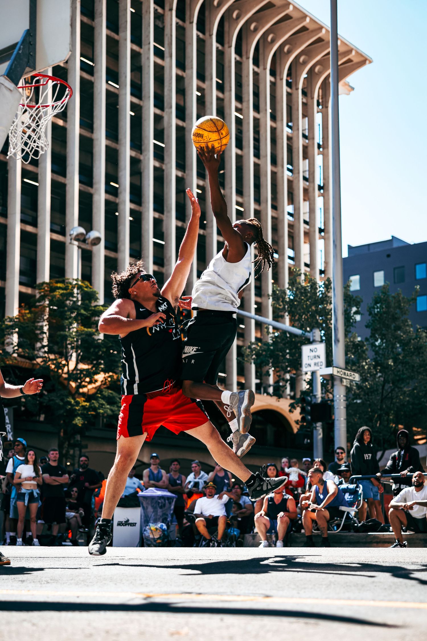 Basketball player in red jersey performs a dynamic layup move against a defender near outdoor columns.