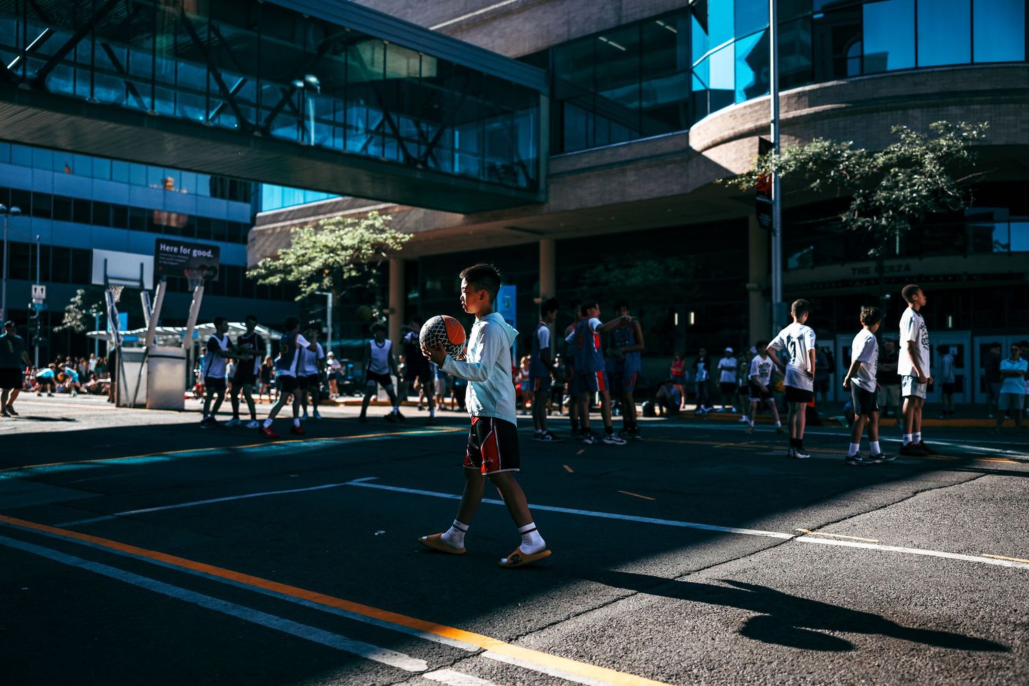People walk through a modern urban plaza with glass buildings and geometric shadows creating dynamic patterns on the ground.
