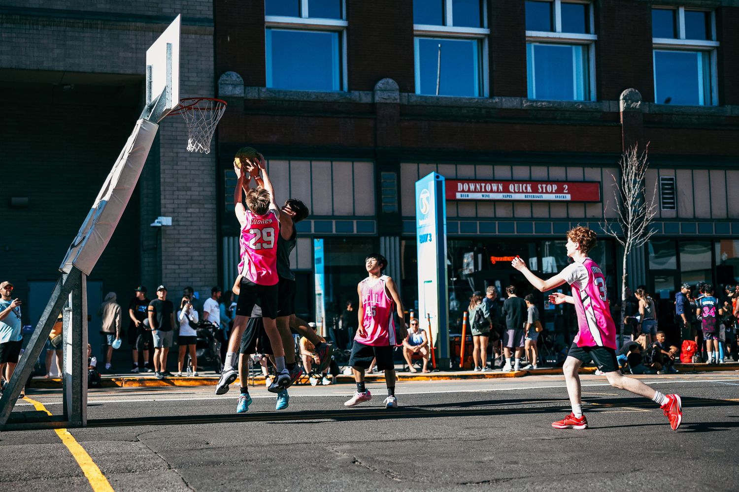 Basketball players compete on an outdoor court during a sunny day with spectators watching from the sidelines.