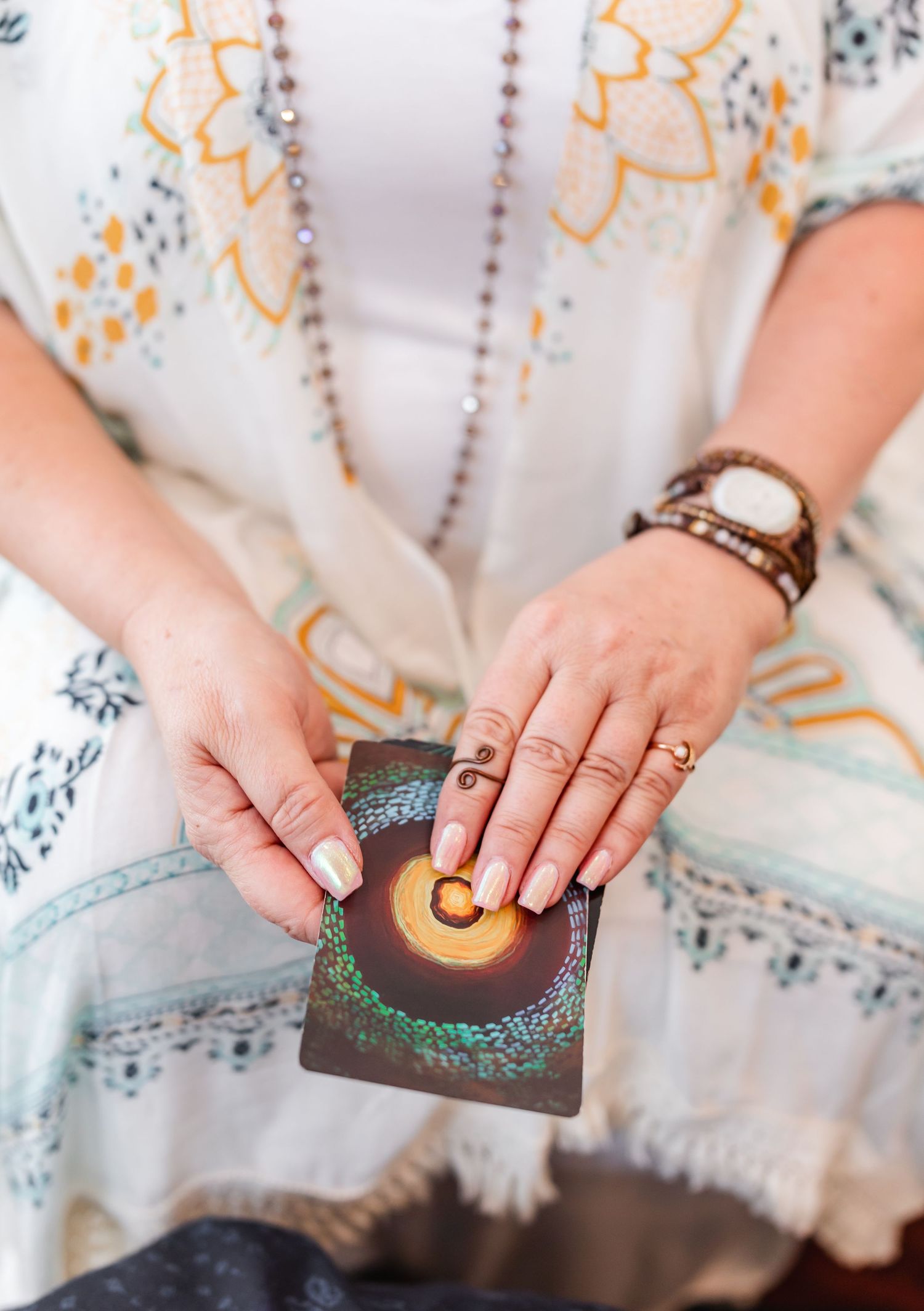 Close-up of a woman’s hands holding an oracle card with a circular design. She wears beaded bracelets, rings, and a white patterned robe wit