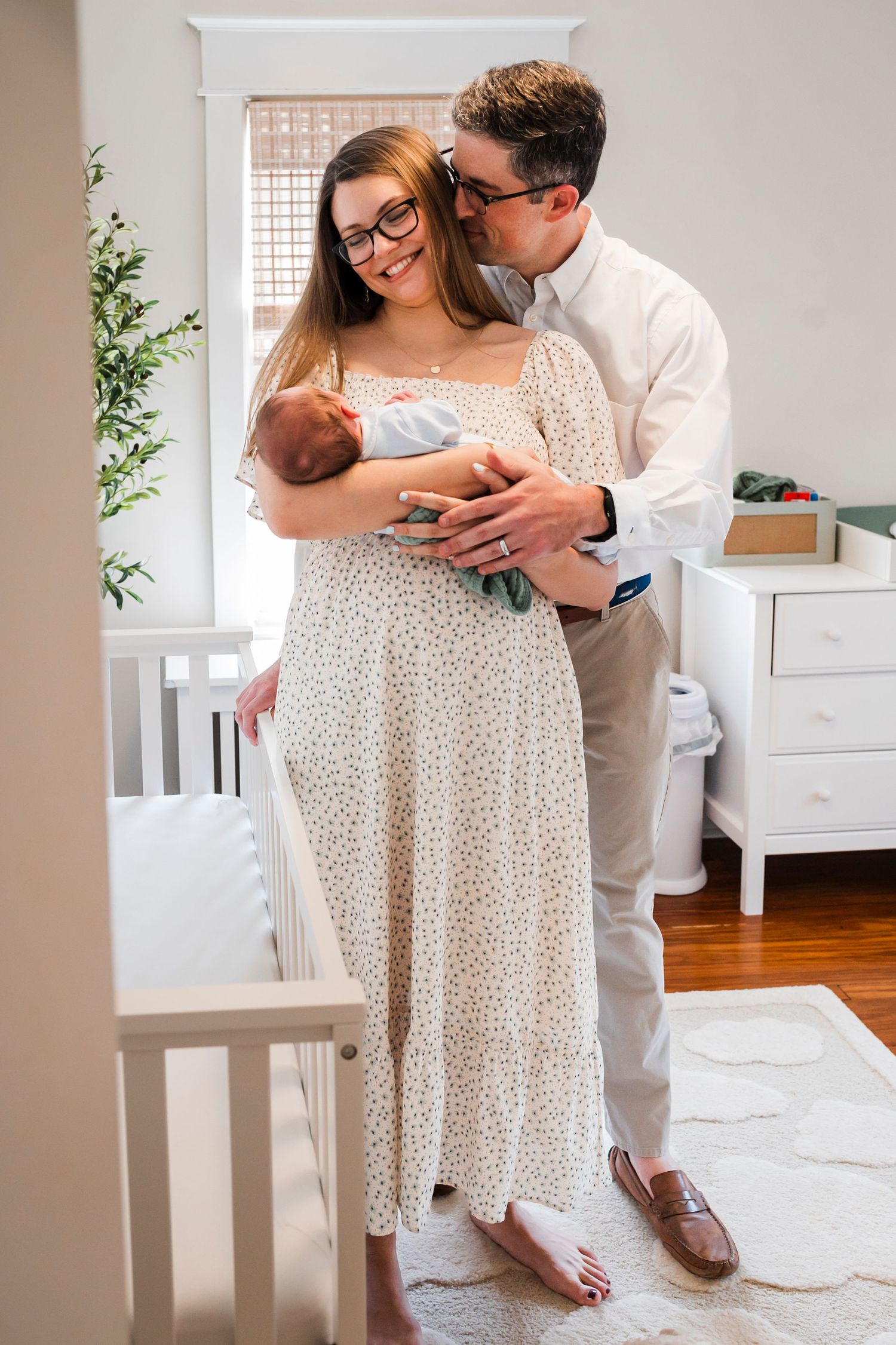 A couple in white outfits share an intimate moment in a bright bedroom with a white crib and furniture.