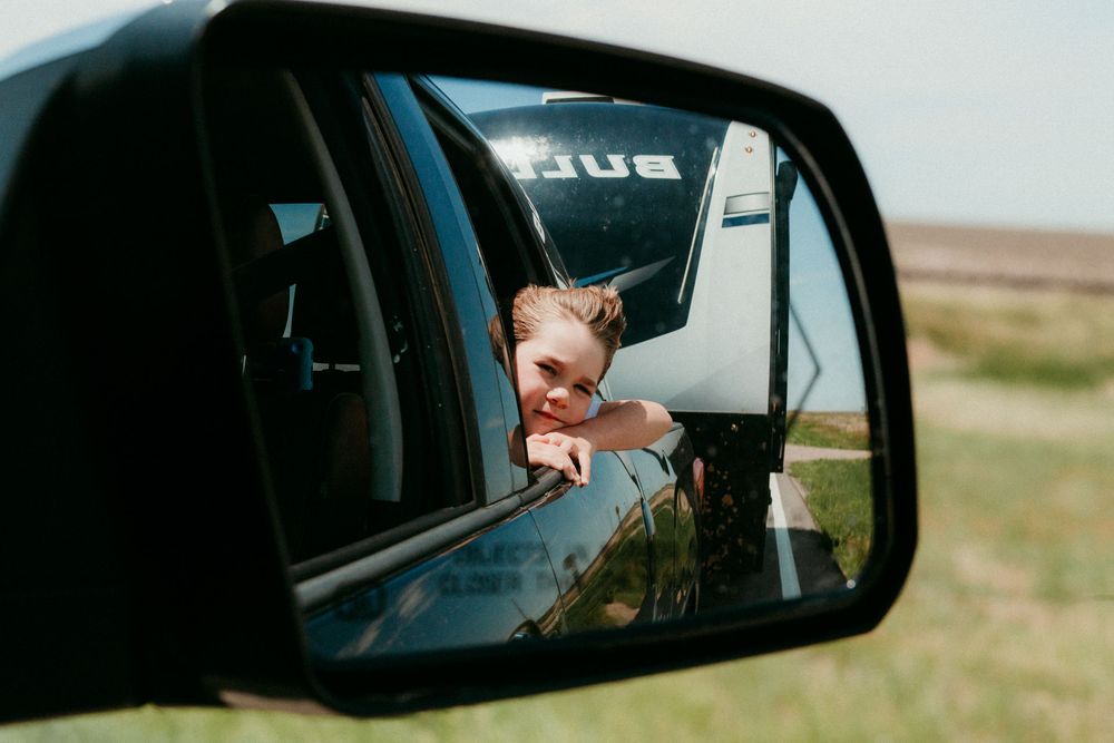 Side view mirror of a car reflects a passenger leaning out the window against a rural landscape backdrop.