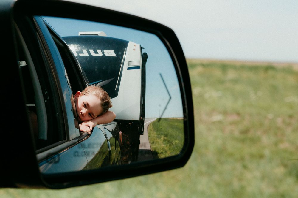 A car side mirror reflects a view of someone looking out the window with a green field in the background.