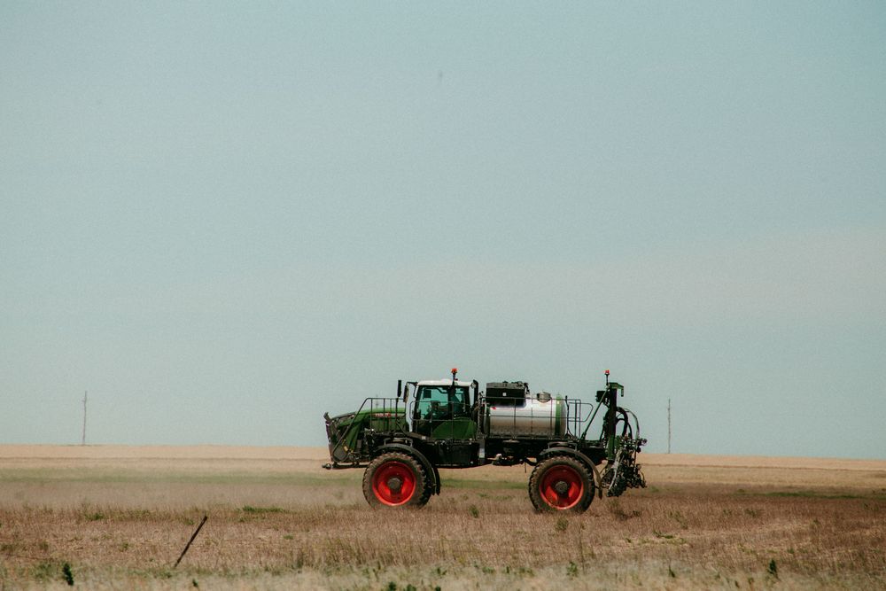 A green tractor drives across a vast golden wheat field during harvest season, kicking up dust in its path.
