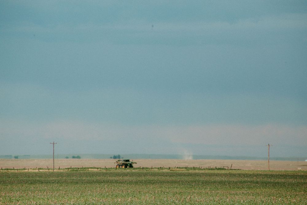 A lone tractor works in a vast agricultural field beneath a turquoise sky on the rural prairie landscape.