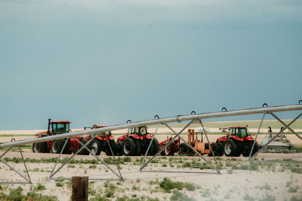 Red tractors parked in a row behind irrigation equipment on a dusty farm field against pale blue sky.