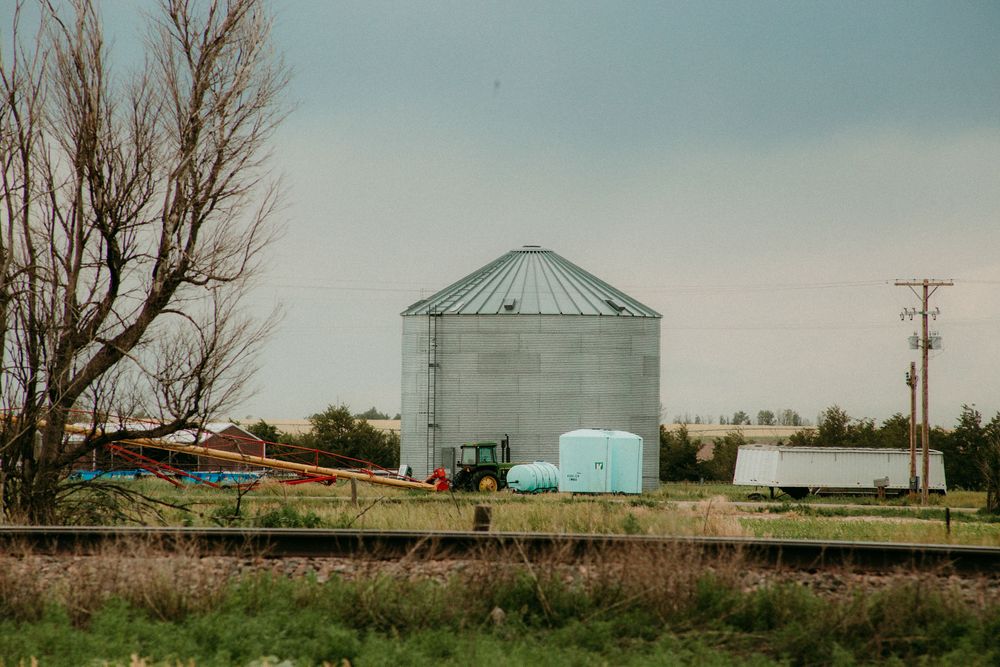 A metal grain silo stands in a rural landscape with farm equipment and a trailer visible nearby on a cloudy day.