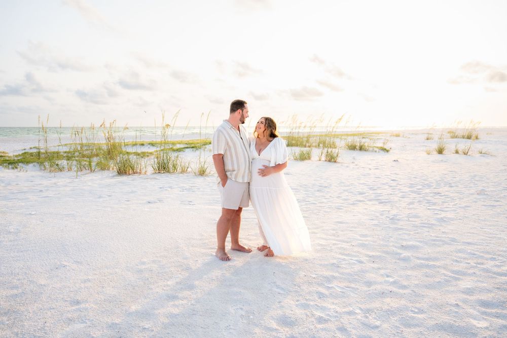 Maternity couple gazing at each other on a serene white sand beach.