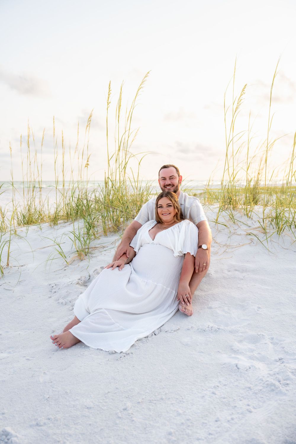 Couple sitting on white sand beach dunes, woman in white maternity dress leaning against man at sunset.