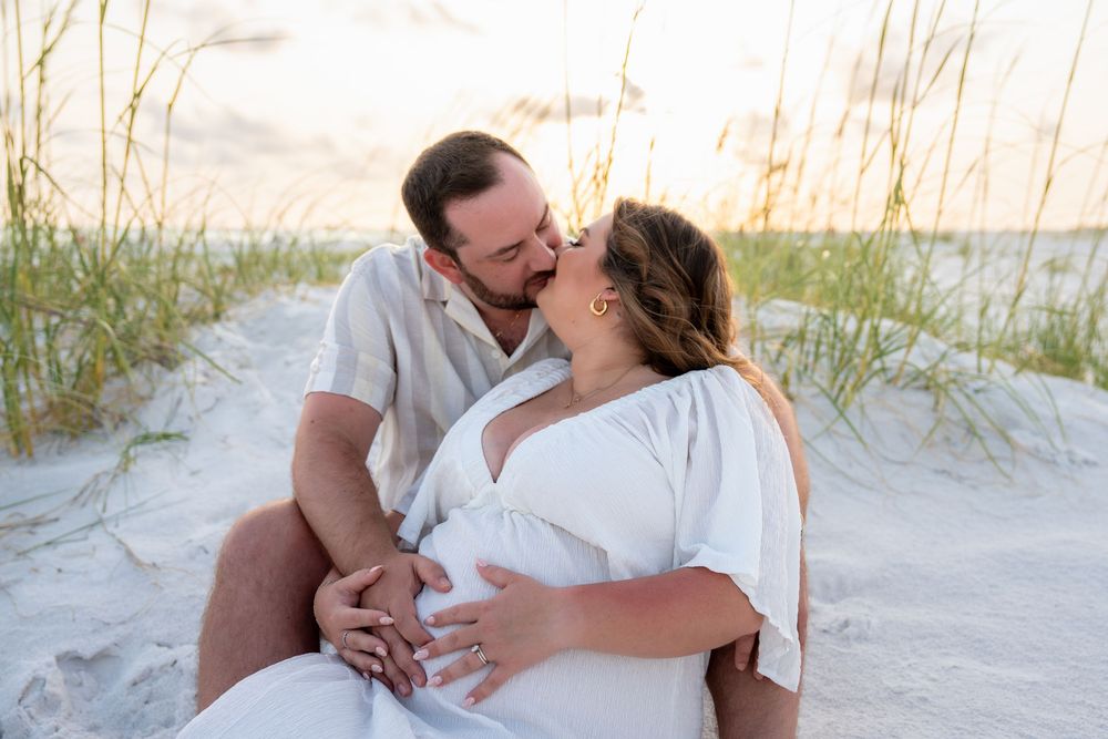 Couple sharing a kiss on white sand beach dunes, hands cradling pregnant belly at sunset.