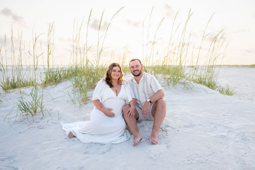 Smiling couple sitting on white sand beach dunes at sunset, pregnant woman in flowing white dress.