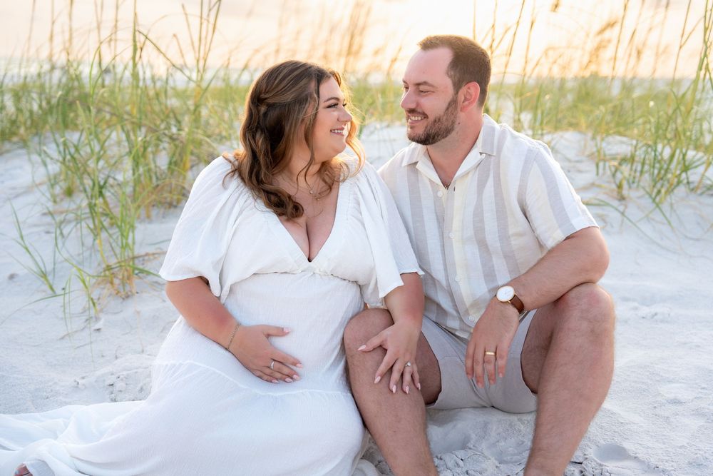 Couple holding hands and smiling at each other on sandy beach, pregnant woman in white maternity dress.