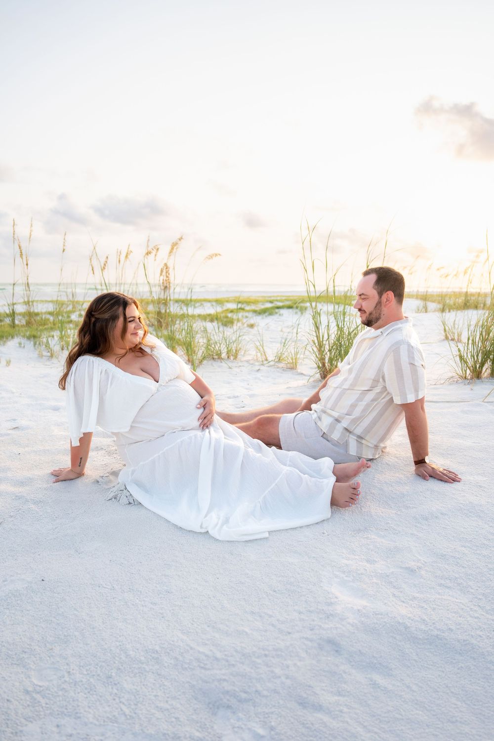 Couple relaxing on white sand beach, pregnant woman in white dress laughing with partner at sunset.