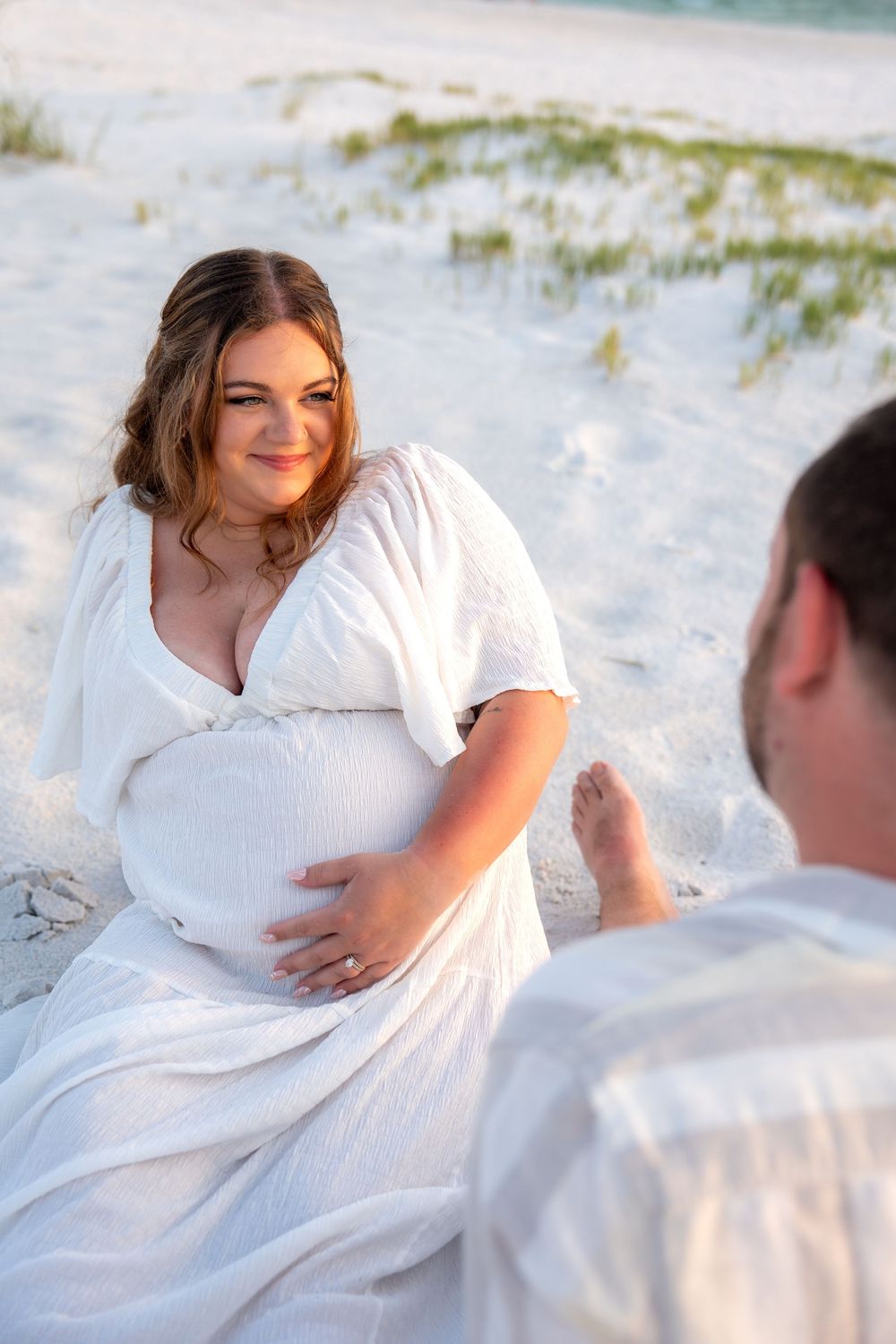 Pregnant woman in white dress sits on sandy beach smiling at partner during golden hour maternity photoshoot.