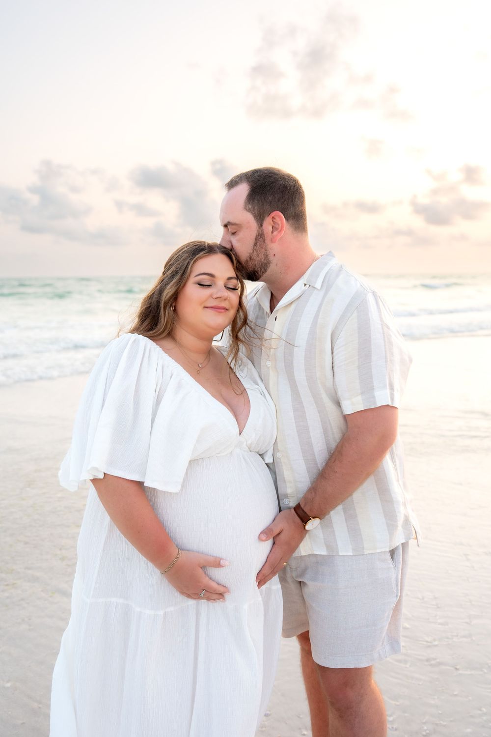 Tender moment as man kisses pregnant partners head while holding her bump on beach at sunset.